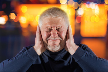 An elderly man suffers from strong noises. He covers his ears with his hands.