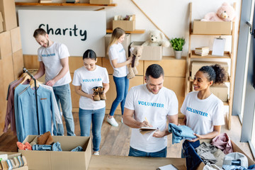 young, cheerful multicultural volunteers in white t-shirts with volunteer inscriptions working in...
