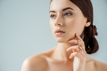 young woman with shiny makeup and golden rings touching face and looking away isolated on grey