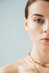 cropped view of young woman with shiny makeup in golden necklaces looking at camera isolated on grey