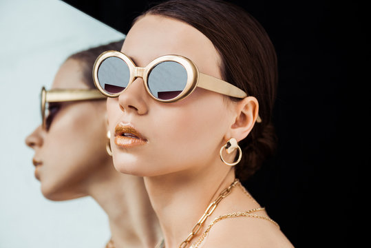 young naked woman in sunglasses, golden jewelry near mirror isolated on black