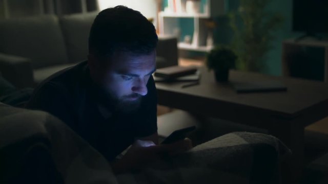Young man watching movie on smartphone lying on sofa at home at night