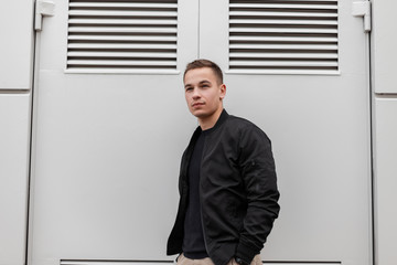 Modern handsome nice young man in fashionable black clothes is standing near metal gray doors to...