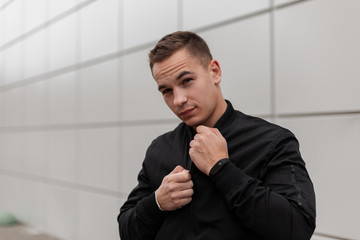 Cute handsome young man in a fashionable black jacket posing standing near a modern building...