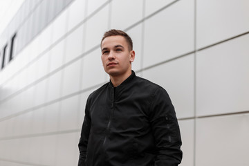 Modern cute young man with a stylish hairstyle in a spring black trendy jacket stands near a gray vintage building outdoors in the city. Handsome european guy. © alones