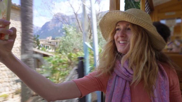 A young woman wearing straw hat enjoying traveling on an old tram or train taking a selfie with beautiful tourist locations using smartphone feeling excited and happy