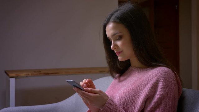 Closeup shoot of young pretty brunette caucasian female texting on the phone and laughing sitting on the couch in a cozy apartment indoors