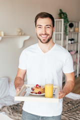 Fototapeta na wymiar smiling man holding tray with pancakes and glass of orange juice in bedroom