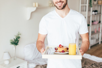 cropped view of smiling man holding tray with pancakes and glass of orange juice in bedroom