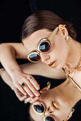 young nude woman in sunglasses, golden jewelry lying on mirror isolated on black