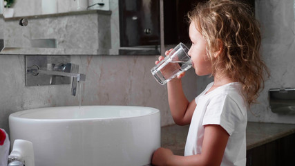 Cute little curly child girl rinses her mouth with water, looking at mirror and spits water into...