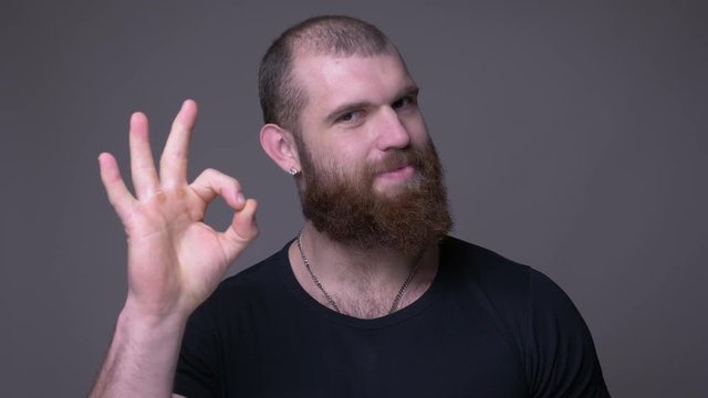 Closeup shoot of adult handsome muscular caucasian man with beard showing okay handsign smiling and looking at camera with background isolated on gray