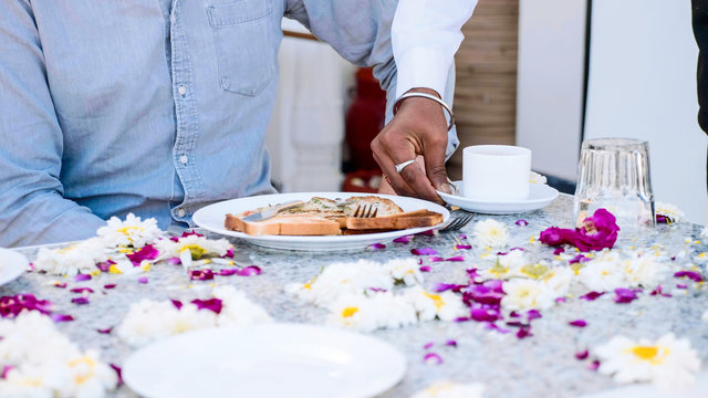 Waiter brings breakfast toasts and pancakes to a couple sitting at the table covered with flowers petals in hotel. Table close-up.
