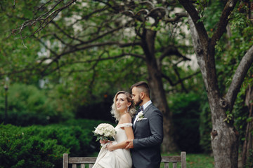 Beautiful bride in a long white dress. Handsome groom in a black suit. Couple in a summer park sitting on a bench