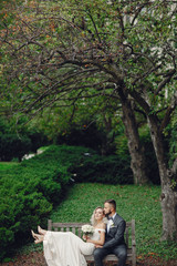 Fototapeta premium Beautiful bride in a long white dress. Handsome groom in a black suit. Couple in a summer park sitting on a bench