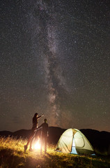 Silhouette of young woman with son in summer camping in the mountains, standing beside campfire and illuminated tourist tent. Mother pointing at night starry sky full of stars and Milky way. Back view