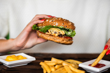 Woman's hand dipping french fries in barbecue sauce. Delicious fresh burger with tomato and cheese. Fast food concept. Beef burger in hands with french fries on rustic wooden board.