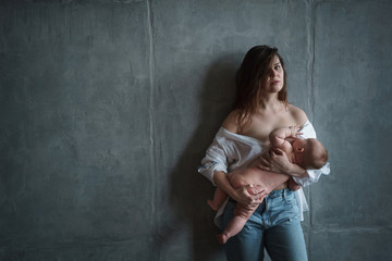 A young mother with a pensive look in white shirt, lowered on her shoulders, and jeans stands at the gray concrete wall holds the baby in her hands and breastfeeds.