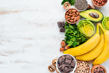 Foods containing natural magnesium. Mg: Chocolate, banana, cocoa, nuts, avocados, broccoli, almonds. Top view. On a white wooden background.
