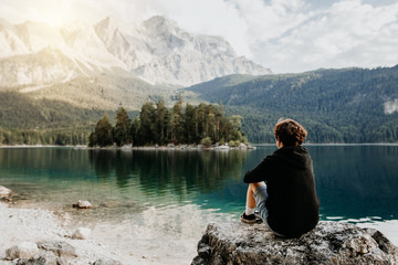 Mensch sitzt auf einem Felsen mit Blick auf einen wunderschönen See mit Bergen und Bäumen im Hintergrund