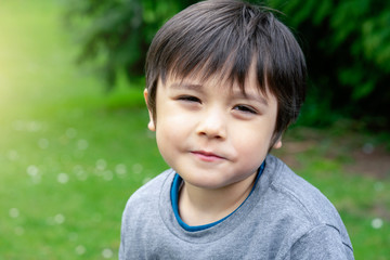 Head shot of happy kid sitting in the park with bright light sunny day, Portrait child playing outdoor in summer with blurry green natural park background, Positive children concept