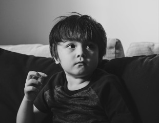 Black and white photo of kid boy sitting on sofa and looking up with thinking face, High key light Portrait of Upset little boy sitting alone, Toddler sad face Dramatic shot of Unhappy child