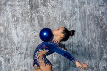 Girl gymnast in a blue suit makes exercise with a ball against a gray wall. Half-length portrait.