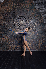 Girl gymnast in a blue suit with sparkles doing exercise with gymnastic clubs is standing on the background of brown wall