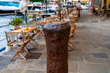 Rusty bollard on the background of a blurred image of a cozy street café and luxury yachts moored at Portofino harbor. Travel and vacation concept.