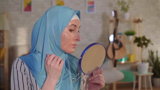 Muslim woman in a hijab looks in the mirror at her scar from a burn on her face