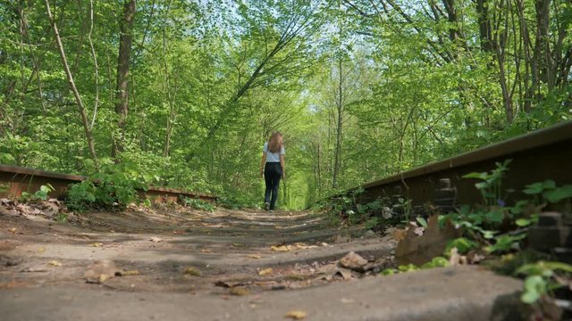 a girl in black pants walks along cross ties and rails against a background of a tunnel of bushes and trees.