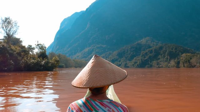 Woman with traditional hat cruising on the brown water of the Nam Ou river in Laos, amazing landscape mountain jungle famous travel destination in South East Asia rear view