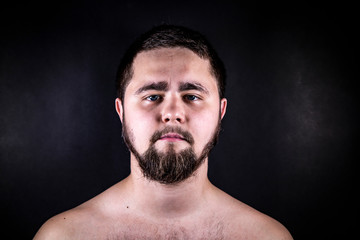 A young man stands with a bare torso on a black background. A man with a beard posing on the camera.