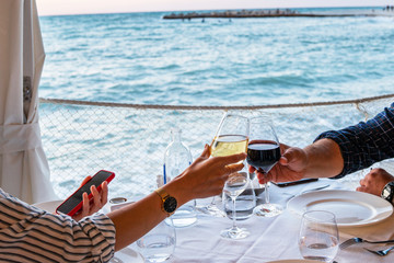 Cheers with wine glasses in a beautiful sunset beach setting. Happy loving couple enjoying dinner in a restaurant. Man and woman enjoying a glass of wine outdoors. Love, dating, food, lifestyle.