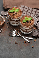 two portions Classic tiramisu dessert in a glass and chocolate bar on dark concrete background