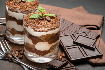 two portions Classic tiramisu dessert in a glass and chocolate bar on dark concrete background