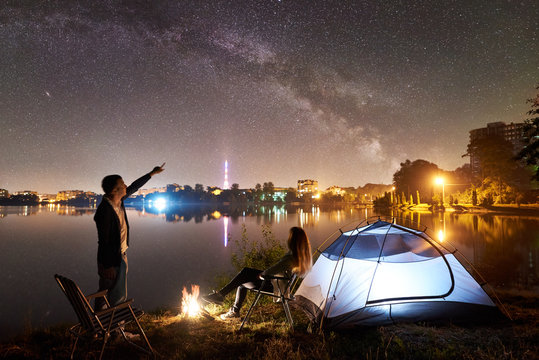 Night camping on lake shore. Couple tourists having a rest near tent and campfire. Man pointing to beautiful night sky full of stars and Milky way, city lights on background. Outdoor lifestyle concept