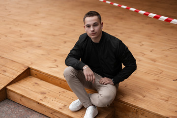Urban young man in stylish sneakers in vintage pants in trendy black jacket sits on a wooden...