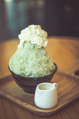 Bingsoo traditional ingredients are, shaved ice, ice cream, in this black cup are green tea bingsoo with whipped cream on top and have condensed milk in a cup ceramic all placed on a wooden tray.