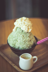 Bingsoo traditional ingredients are, shaved ice, ice cream, in this black cup are green tea bingsoo with whipped cream on top and have condensed milk in a cup ceramic all placed on a wooden tray.