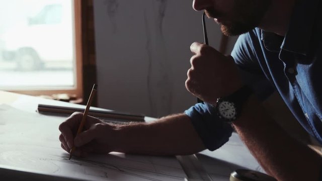 Profile of attractive professional painter holding glasses drawing with pencil inside a wooden workshop