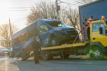 Loading a broken blue minibus on a yellow tow truck in the middle of the street on a sunny day