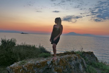 girl stands on a rock and looks at the beautiful view of the sea and sunset