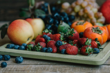 Healthy food clean eating selection in wooden tray: strawberries, blueberries apples, grapes, persimmon, pomegranate on rustic wooden background. Toned image.