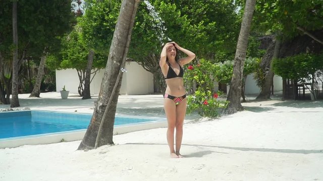 Young brunette caucasian woman in swimsuit takes refreshing tropical shower in luxury resort garden on beach amidst green foliage, palm trees and bushes on bright hot sunny day. Slow motion footage.