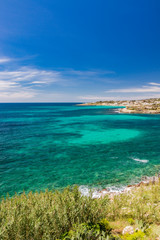 Fototapeta na wymiar Spectacular view of the Apulian coast in Leuca, in Puglia, Salento, Italy. Turquoise sea, clear blue sky, rocks, sun, wind, white clouds, lush vegetation in summer. Populated area with white houses.