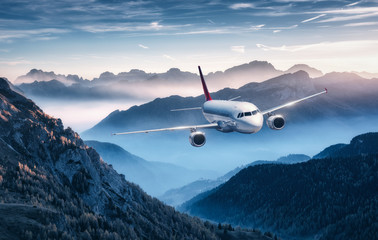 Airplane is flying over mountains in fog at sunset in summer. Landscape with passenger airplane, hills in low clouds, blue sky. White aircraft. Business travel. Commercial plane. Aerial view