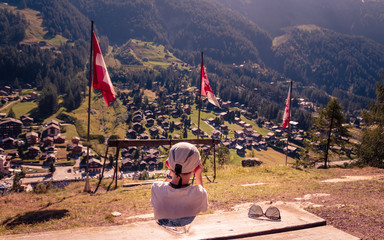 Young boy sitting at lookout point over alpine village Zinal in summer at wooden table with...