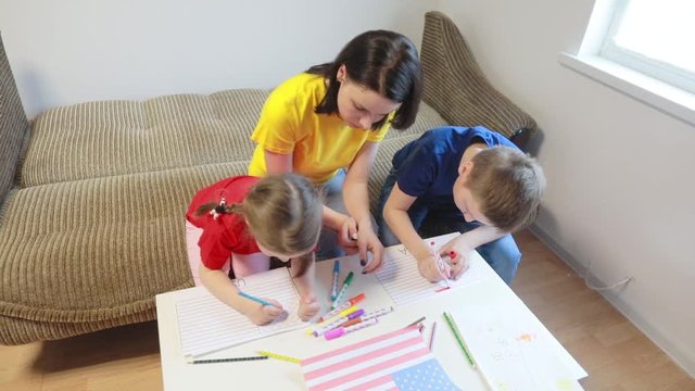 A caring mother teaches two of her children to draw the flag of America with colored pencils and felt-tip pens for America’s Independence Day.