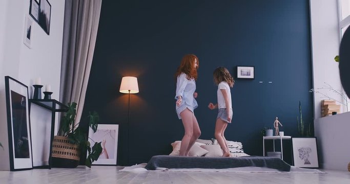 Happy cute daughter and young mother jumping and dancing on bed while have fun during holidays at home